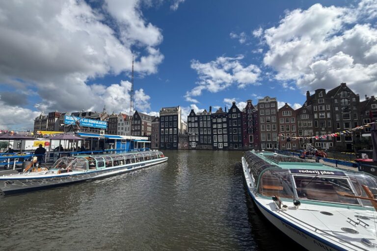 Canal cruise boats docked along the waterfront with historic Dutch canal houses under a dramatic blue sky in central Amsterdam.