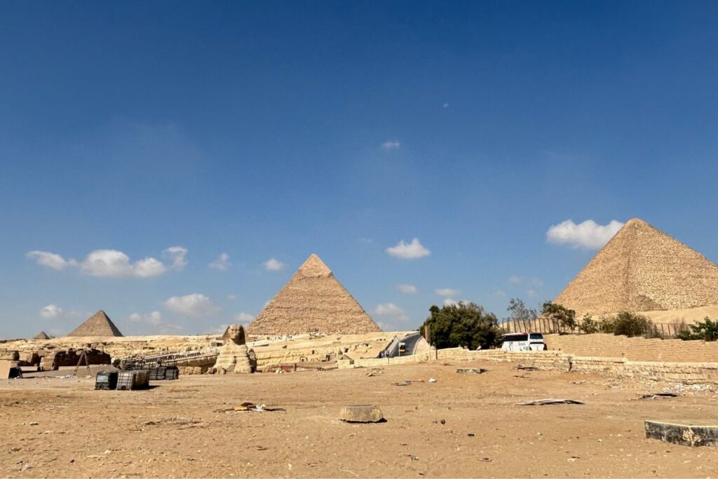 Wide desert view of the pyramids of Giza and the Sphinx beneath a bright blue sky with scattered clouds.