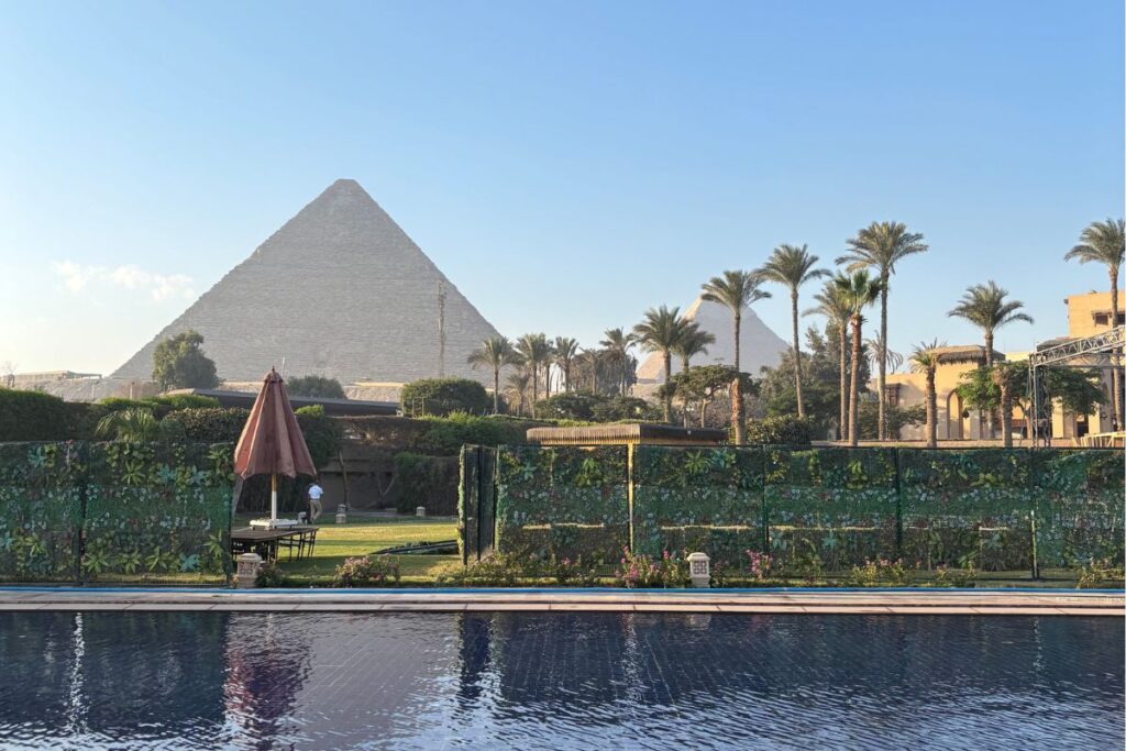 Marriott Mena pool overlooking the pyramids of Giza framed by palm trees and greenery during golden hour.