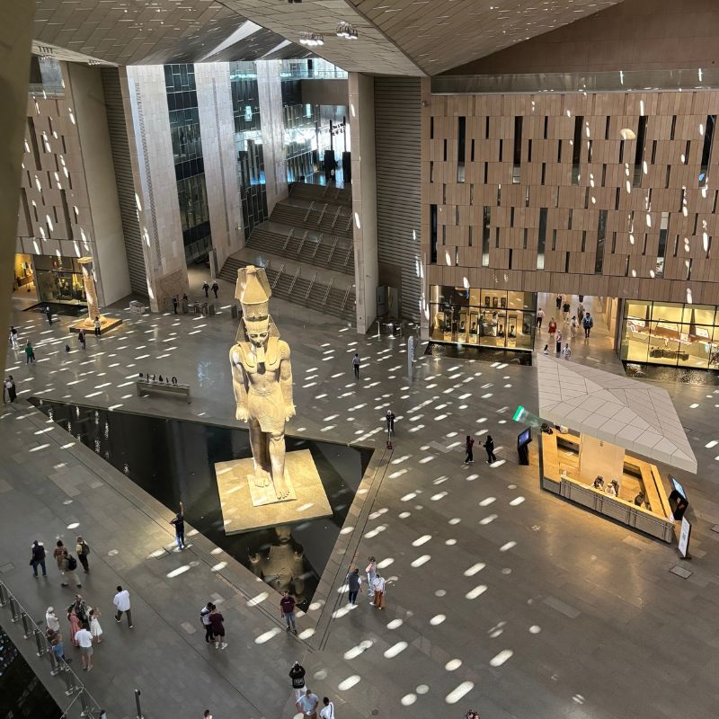 Elevated view inside the Grand Egyptian Museum showing a towering pharaoh statue, reflective pools, and visitors walking through the modern hall.