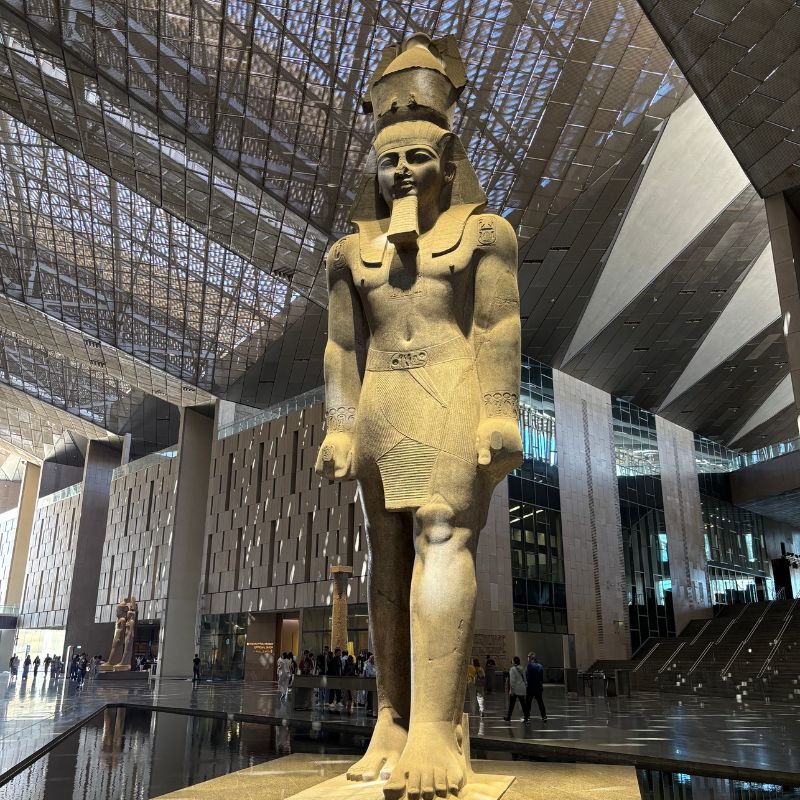 Massive ancient Egyptian statue displayed inside the Grand Egyptian Museum atrium beneath a geometric glass ceiling.