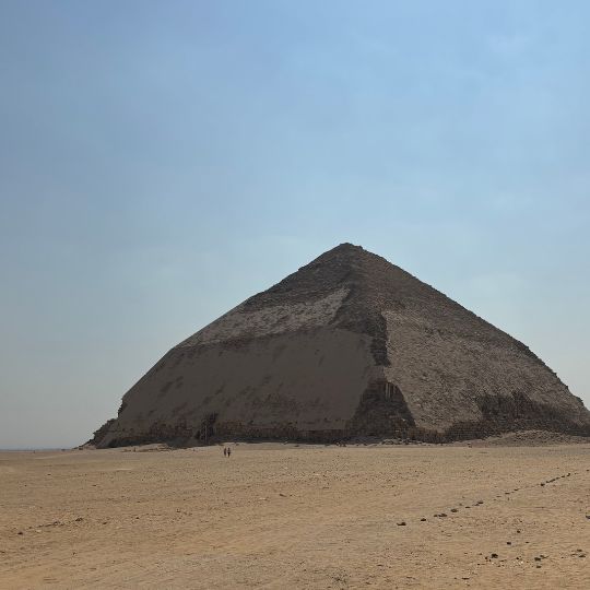 The Bent Pyramid rising from the desert under a pale blue sky with tiny visitors visible near the base.
