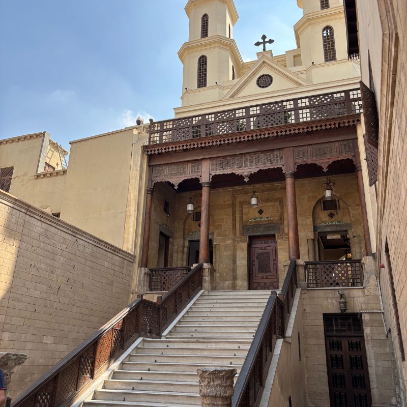 Historic stone staircase leading to a church entrance with carved wooden balconies and cream-colored walls in Old Cairo.