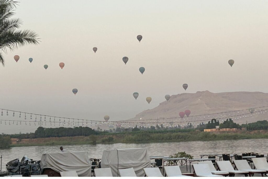 Hot air balloons drifting over the Nile River and desert mountains at sunrise, viewed from a riverside deck at Hilton Luxor Resort.