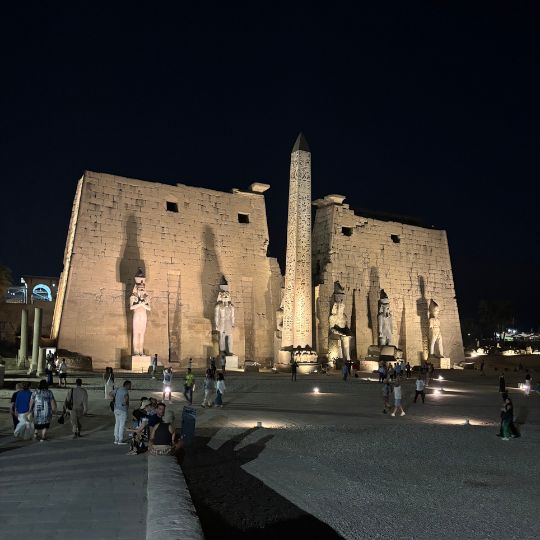 Luxor Temple illuminated at night with towering statues, an obelisk, and visitors walking through the historic complex.