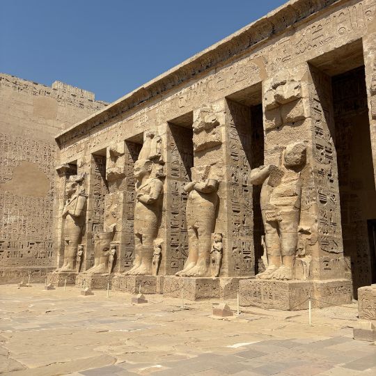 Ancient stone statues lined along the walls of an Egyptian temple courtyard under bright midday sunlight.