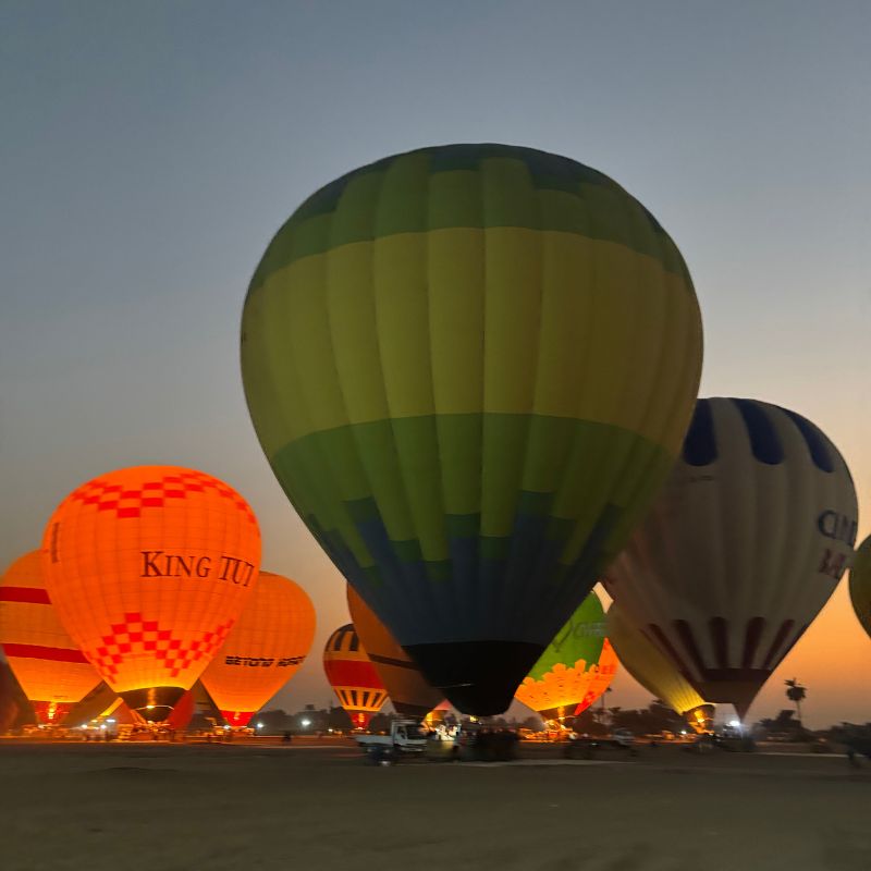 Colorful hot air balloons glowing before sunrise near the pyramids, with dark desert silhouettes in the background.