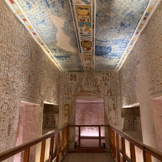 Interior of an ancient Egyptian tomb chamber with colorful hieroglyphics and painted figures covering the walls and ceiling, viewed from a wooden walkway inside the burial site.