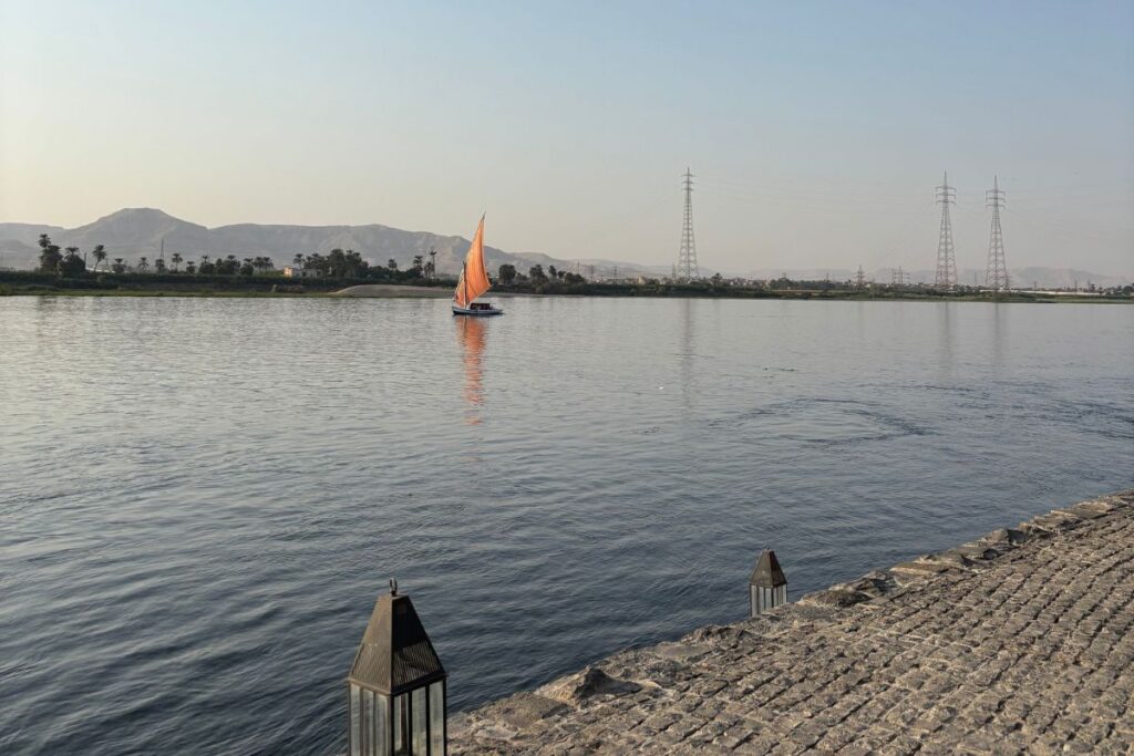Traditional felucca sailboat with orange sail gliding across the Nile River at dusk near Luxor.