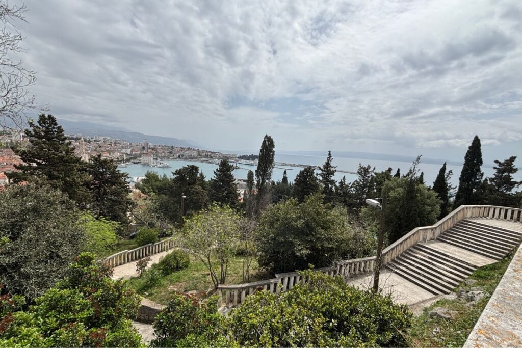 Elevated view over Split’s waterfront and harbor framed by gardens, stone staircases, and cloudy skies.