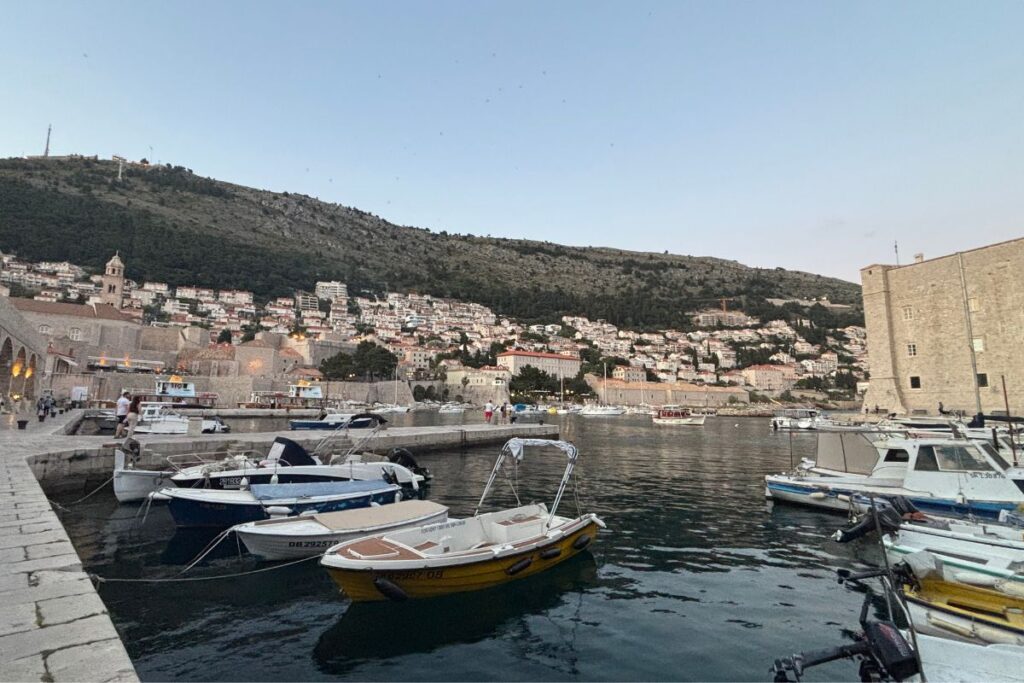 Small boats floating in Dubrovnik’s old harbor at dusk with the hillside city rising behind the waterfront.