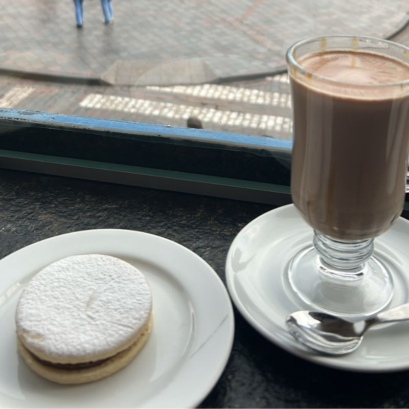 Hot chocolate and a powdered alfajor cookie on a café table overlooking Plaza de Armas through a window.