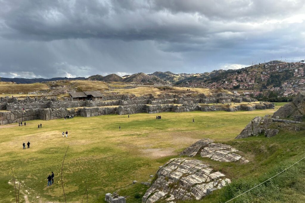 Wide view of Sacsayhuamán with massive Inca stone walls, open grassy fields, and storm clouds gathering over the hills surrounding Cusco.