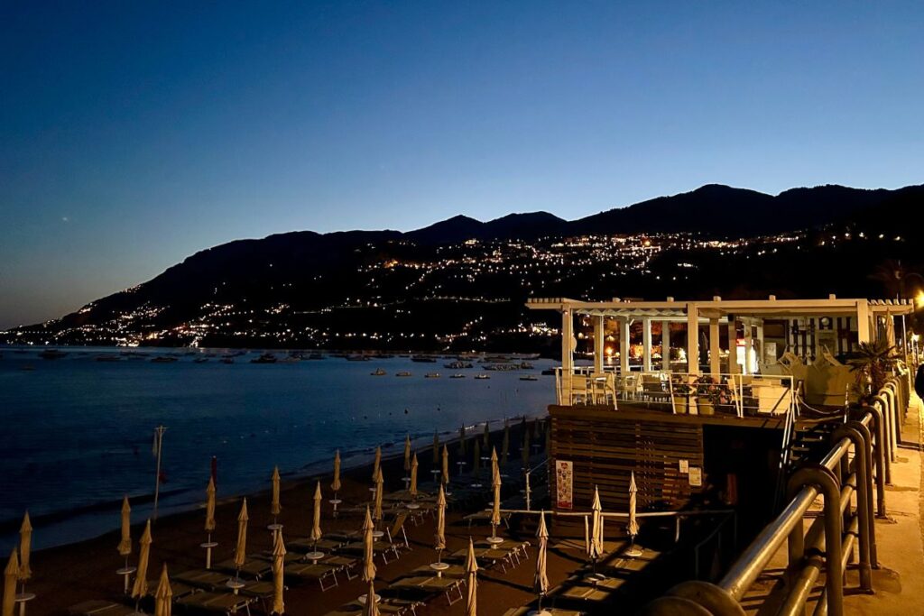 Maiori beach at dusk with rows of closed umbrellas, a waterfront restaurant, and twinkling lights climbing the mountainside along the Amalfi Coast.