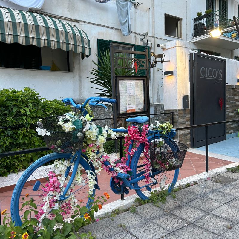 Blue bicycle decorated with colorful flowers outside a restaurant in Maiori, adding a cheerful touch to the seaside town's streets.