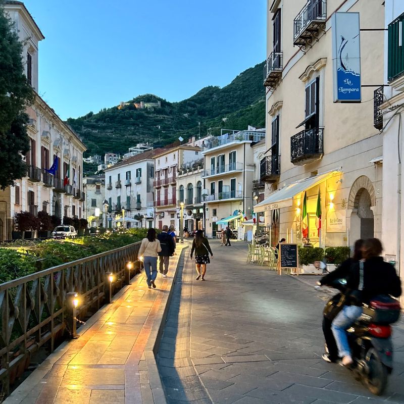Evening stroll through Maiori's town center, with illuminated sidewalks, outdoor cafés, and mountain views rising above the buildings.
