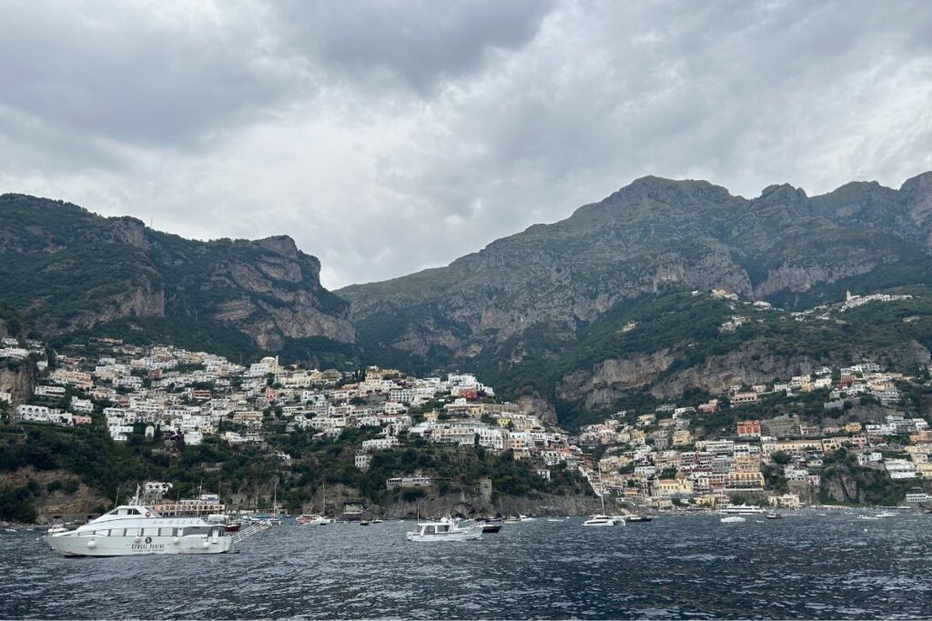 Wide view of Positano and the Amalfi Coast from a ferry, with dramatic mountains, cliffside homes, and boats floating in the bay beneath cloudy skies.