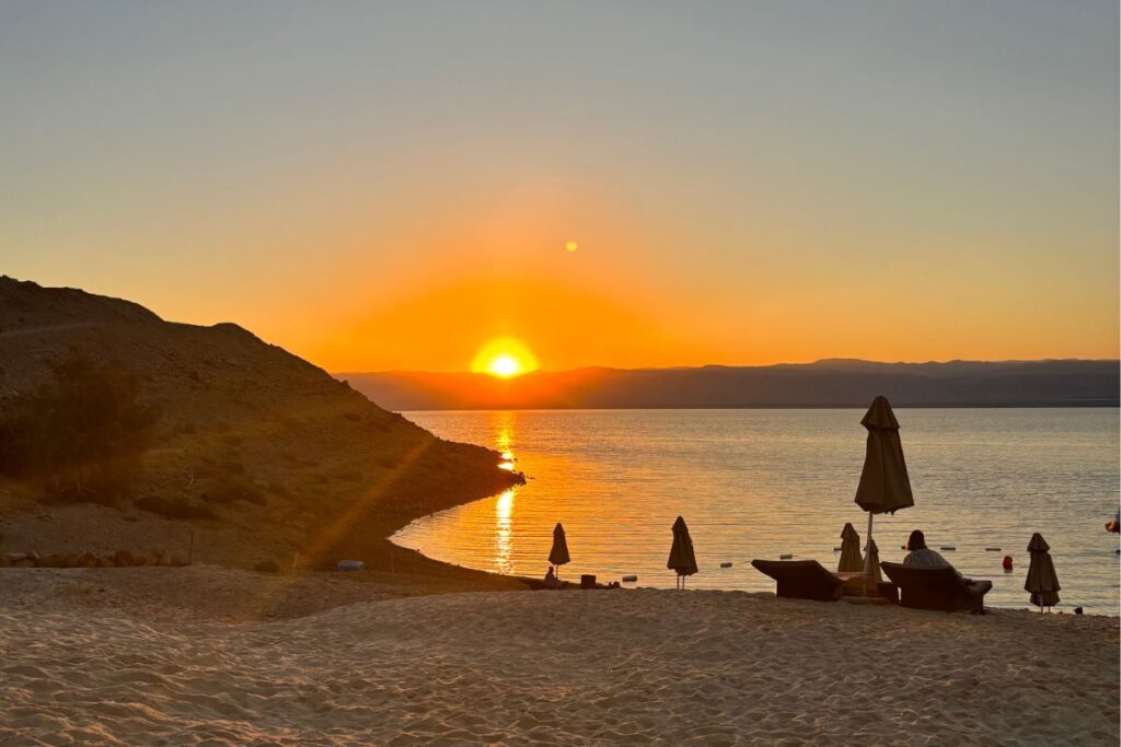 Golden sunset over a quiet beach and calm water, with lounge chairs and umbrellas silhouetted against the horizon. from Hilton Dead Sea Resort