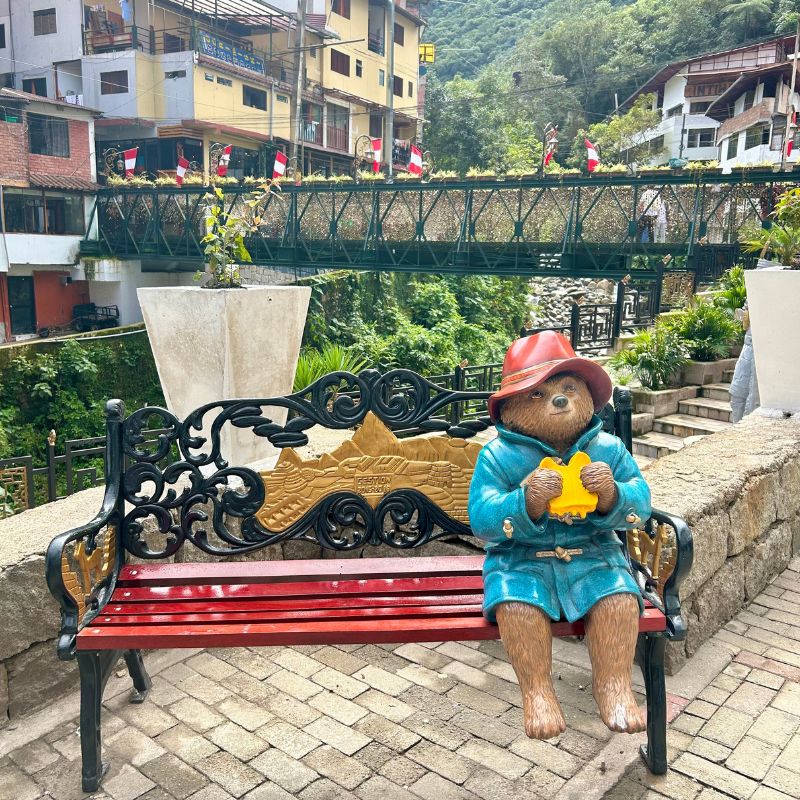Statue of Paddington Bear sitting on a bench in Aguas Calientes with a river bridge and mountain scenery in the background near Machu Picchu.