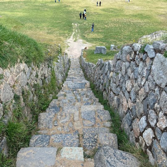 Ancient stone staircase descending through the ruins of Sacsayhuamán overlooking a grassy archaeological site near Cusco.