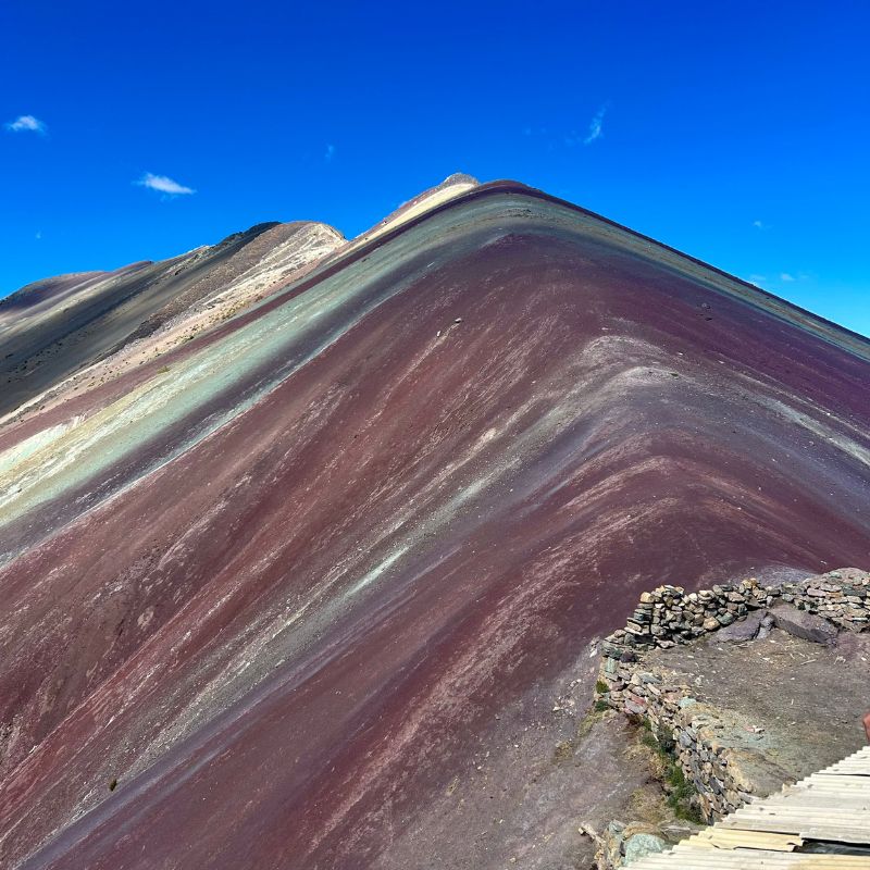 Vibrant striped slopes of Rainbow Mountain under a bright blue sky with wooden steps leading toward the summit viewpoint.