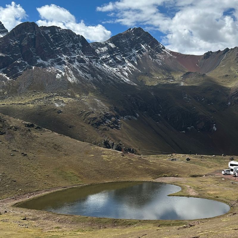 High-altitude Andean landscape near Rainbow Mountain with a small reflective lake surrounded by rugged snow-dusted peaks.