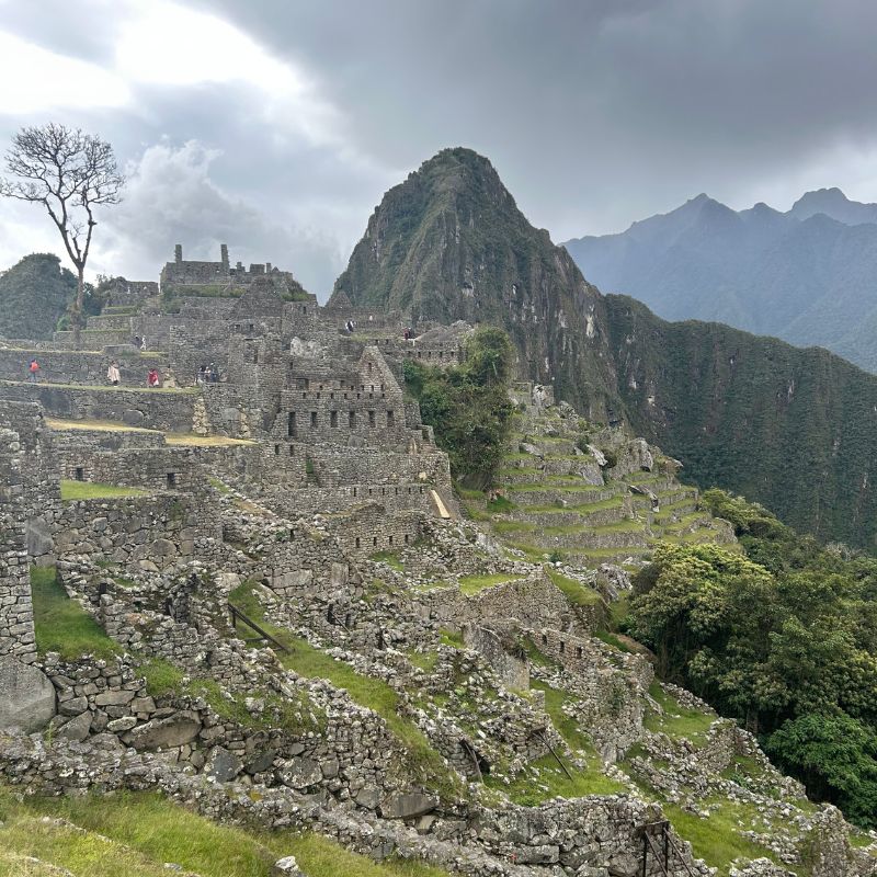 Wide view of the ancient Inca citadel of Machu Picchu under dramatic cloudy skies with terraced stone ruins and surrounding mountain peaks.