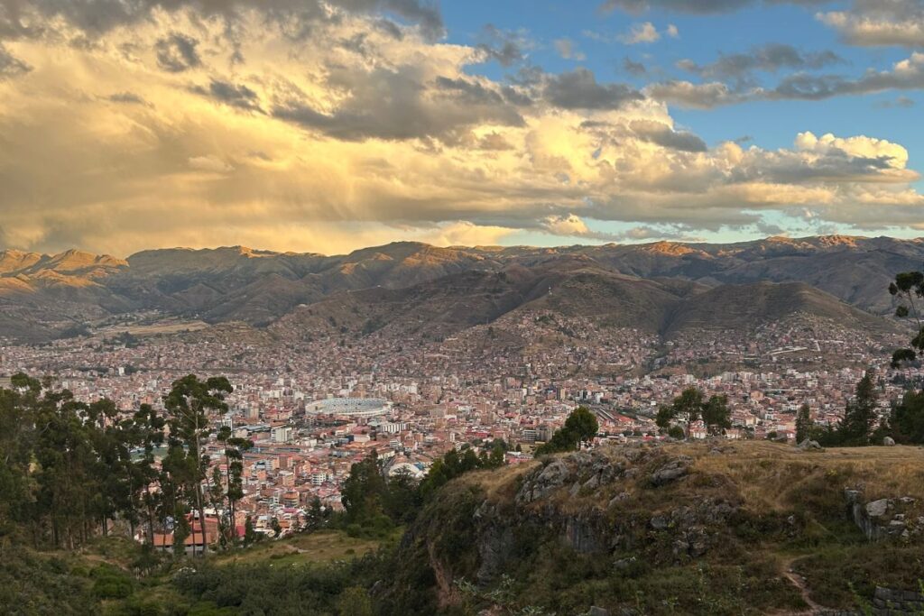 Panoramic sunset view overlooking Cusco with dramatic clouds above the mountains and the city spread across the valley below.