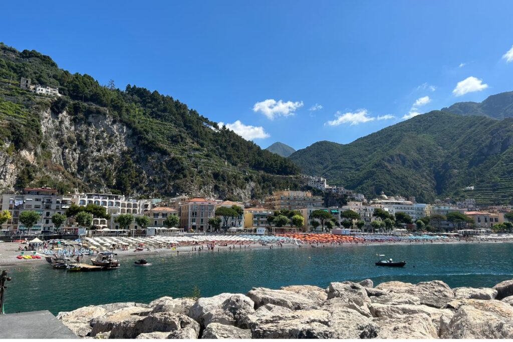 Wide view of Maiori's beach and waterfront, featuring colorful umbrellas, turquoise water, and mountains surrounding the town along the Amalfi Coast.
