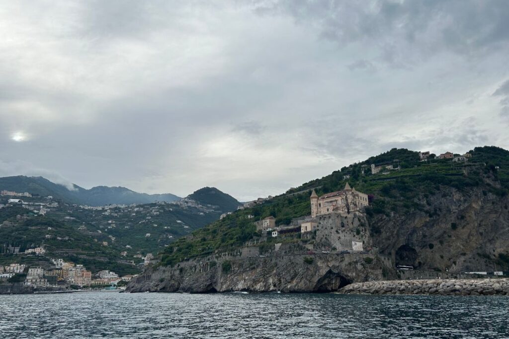 View of the Amalfi Coast from a ferry, with a historic villa perched on rocky cliffs above the sea and terraced hillsides stretching into the distance under a cloudy sky.