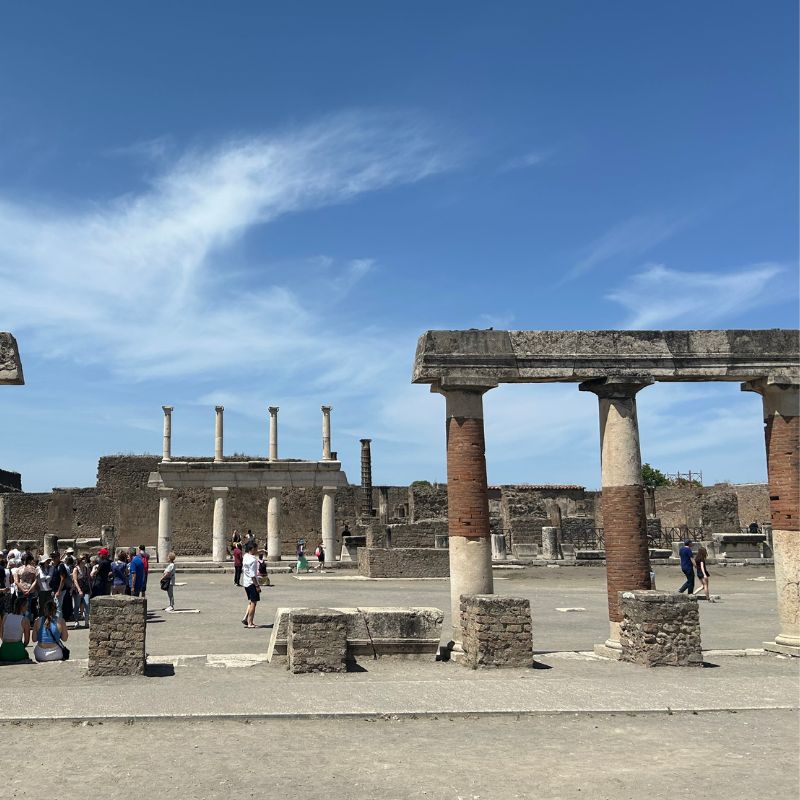 Ancient stone columns and ruins in Pompeii, with visitors exploring the archaeological site beneath a bright blue sky.