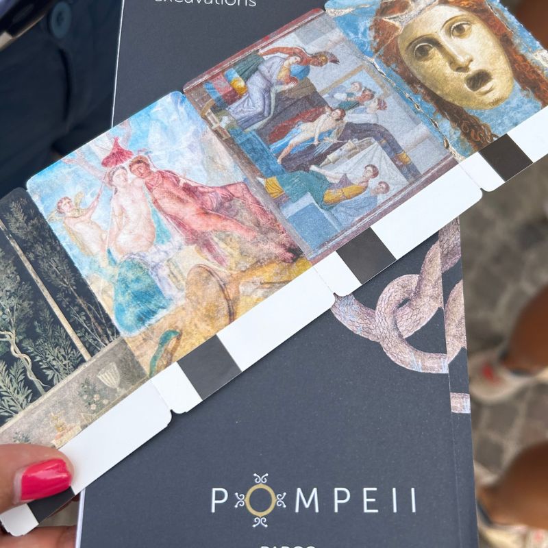 Pompeii admission booklet and illustrated bookmark featuring ancient fresco artwork, held by a visitor during a tour of the archaeological site.