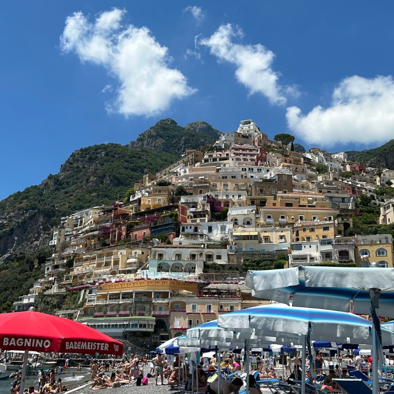 View of Positano's colorful hillside buildings rising above a crowded beach lined with umbrellas along Italy's Amalfi Coast.