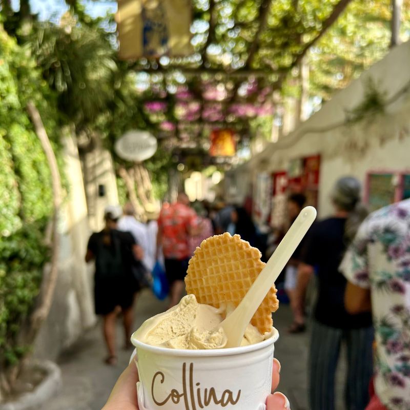 Cup of gelato held along a shaded pedestrian street in Positano, with colorful storefronts and visitors walking through the bustling alleyway behind.