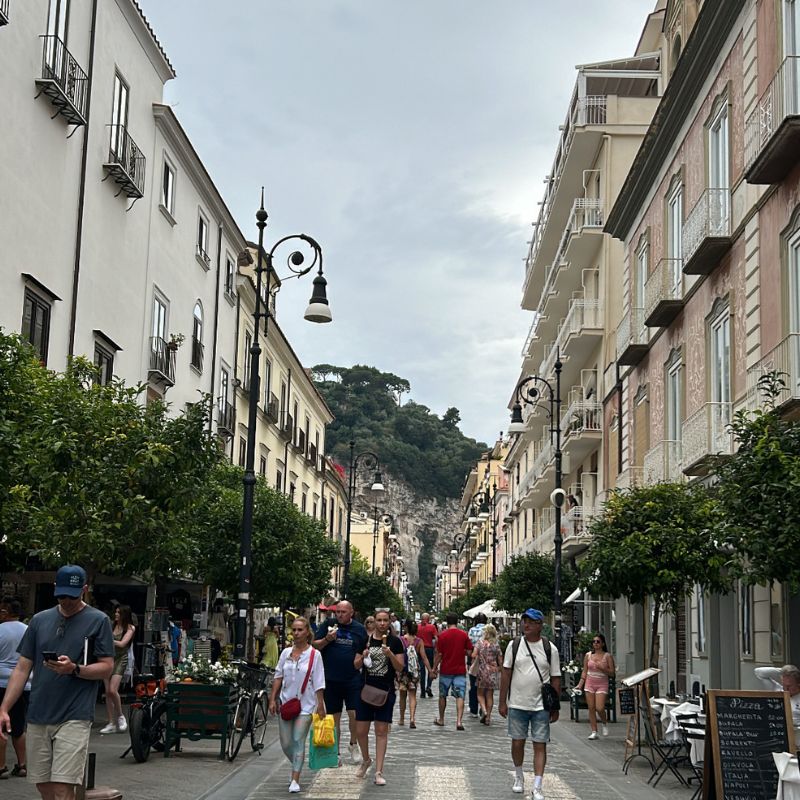 Pedestrian street in Amalfi lined with shops, cafés, and historic buildings, with visitors strolling beneath cloudy skies.