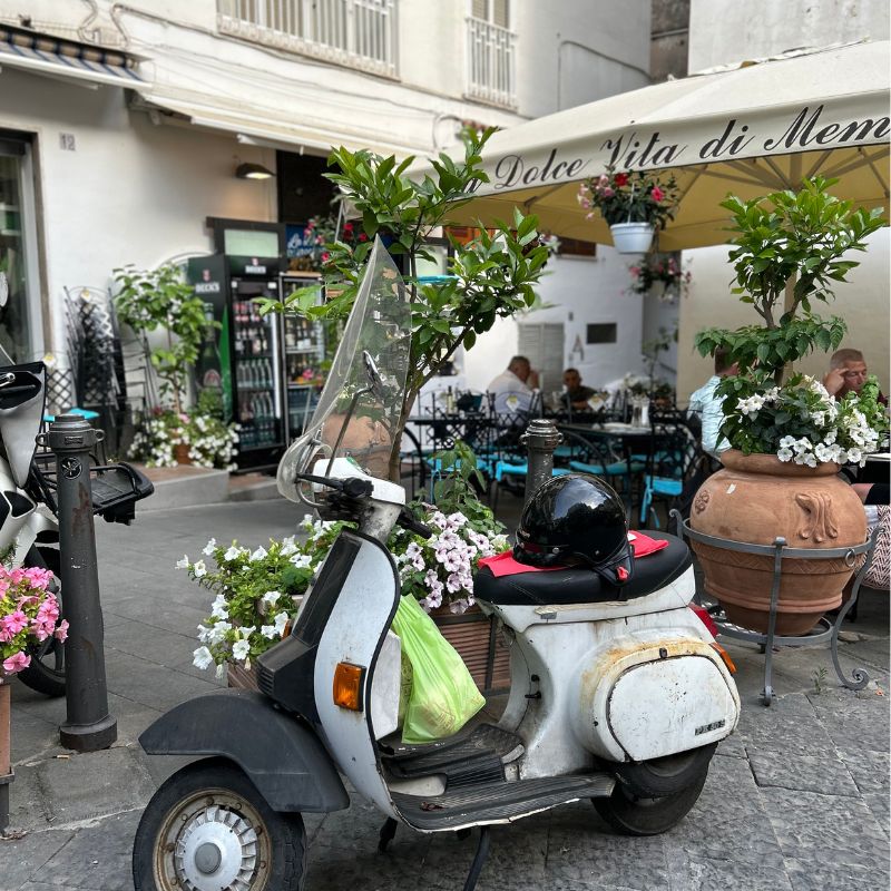 Classic Vespa scooter parked outside a flower-filled café in a charming Amalfi Coast town.