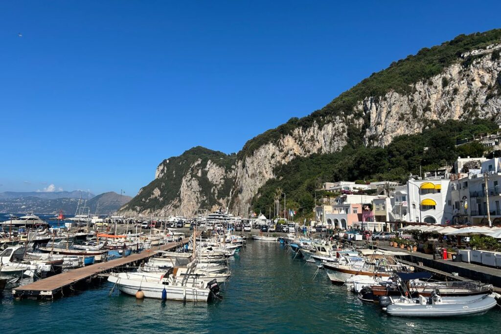 Marina in Capri filled with boats and yachts, framed by colorful waterfront buildings and dramatic limestone cliffs.