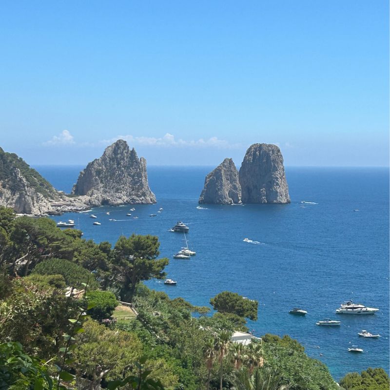 View of Capri's Faraglioni sea stacks rising from the Mediterranean Sea, surrounded by boats and lush coastal vegetation.
