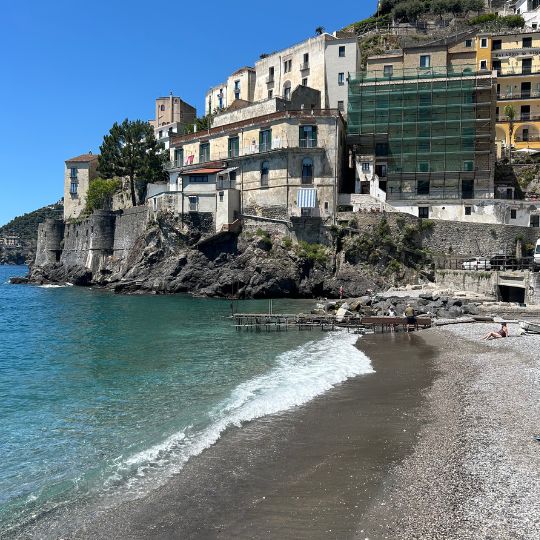 Small beach in Atrani with turquoise water, historic buildings perched on rocky cliffs, and colorful homes overlooking the shoreline.