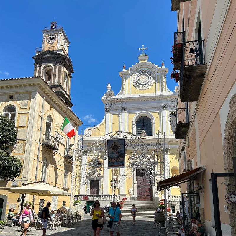 Visitors walking through a sunny piazza in Amalfi, with the cathedral facade and bell tower rising above surrounding cafés and historic buildings.