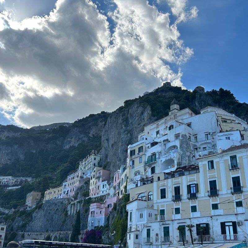 Pastel-colored buildings clinging to the cliffs of Amalfi, with dramatic mountains and clouds towering above the town.