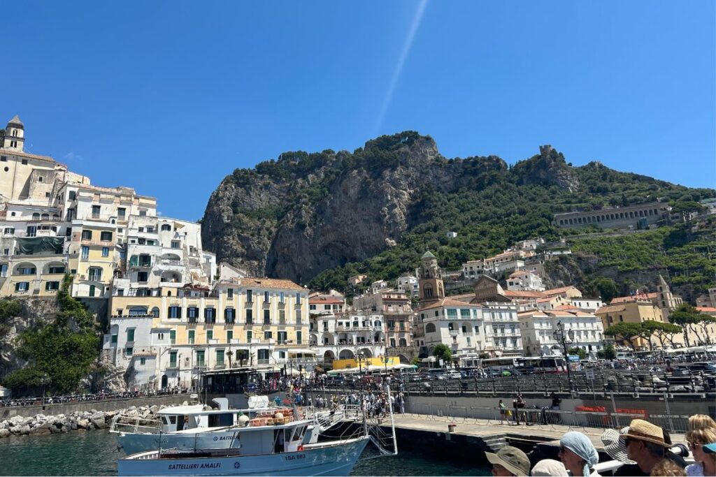 View of Amalfi's waterfront from the harbor, with colorful buildings, fishing boats, and steep cliffs rising behind the town under a clear blue sky.