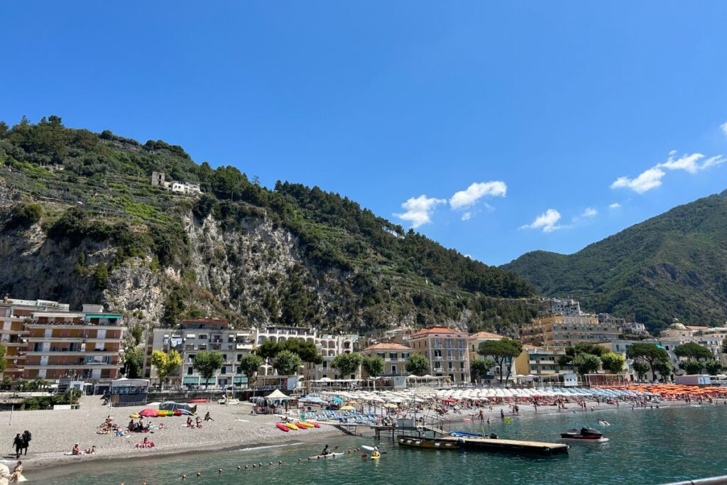 View of Maiori's wide beach and waterfront, with colorful umbrellas, clear water, and mountains rising behind the town on Italy's Amalfi Coast.