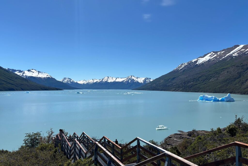 Turquoise glacial lake surrounded by snow-capped mountains and floating icebergs, viewed from a wooden overlook in Patagonia.