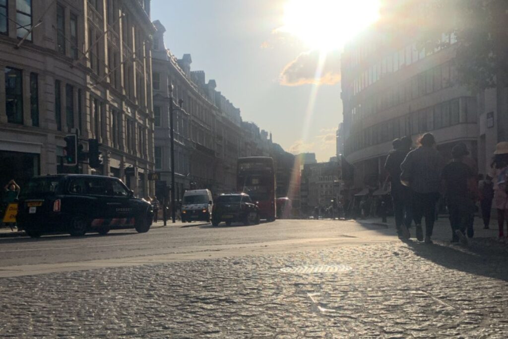 Low-angle view of a busy London street at sunset, with black cabs, pedestrians, and historic buildings silhouetted against the evening light.