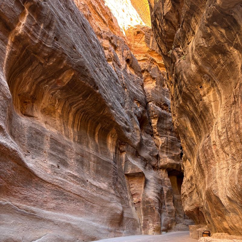 Narrow sandstone canyon walls inside the Siq passage leading toward Petra, with layered rock textures glowing in warm light.