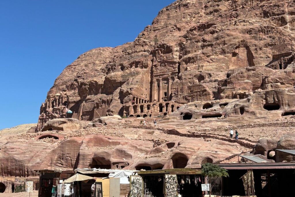 Ancient carved tombs and rock-cut facades built into the sandstone cliffs of Petra under a bright blue sky.