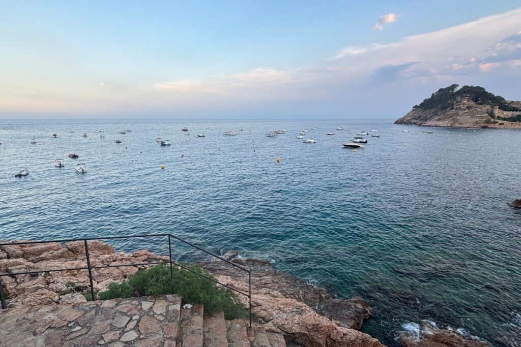 Rocky shoreline and calm Mediterranean waters with small boats anchored off the coast of Tossa de Mar at sunset