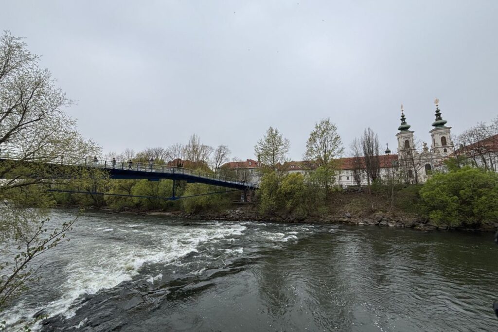 Mur River flowing through Graz with a pedestrian bridge and historic church in the background