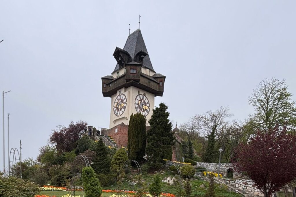 The historic Graz Clock Tower (Uhrturm) atop Schlossberg Hill in Graz, Austria, surrounded by gardens on a cloudy spring day.